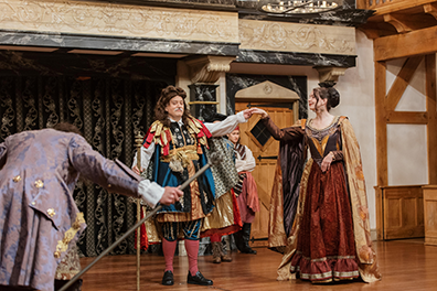 ASC-Two_Gentlemen_of_Verona-141 The Duke in the center of the stage with long curly brown wig, a colorful mesh of robes, a staff in his right hand and his left hand holding high the and of Silvia in a burgundy and gold Jacobean dress and gold cape as, in the front left corner, Valentine bows, his light purple coat's back to us.