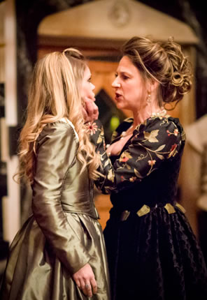 Livia snarling in the face of and her left hand gripping the chin of Isabella, who is wearing a classic greenish gold Elizabethan dress