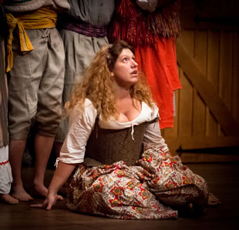 The Jailer's Daughter, in country patterned Elizabethan dress, kneels on the floor looking up in distraction with the legs of the Morris dancers behind her.