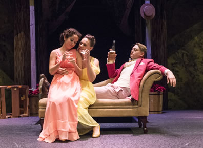 Hermia in a peach gown sits on the lap of Helena in a yellow gown, who sits on the edge of the chaise lounge as Lysander in salmon jacket leans back on the chaise lounge. The women have champagne glasses, Lysander the bottle.