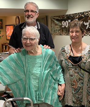 Carol, Eric, and Sarah Carol Kelly in a green blouse and house jacket holding hands with Sarah in a colorful tapestry dress and me in black polo shirt with my hands on Carol's shoulders. In the background is a 90th Birthday! banner