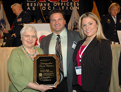 Carol_Matt_Julie Carol holds the ROA Distinguished Service Citation plaque in her hands as she stands next to Matt, in tie and jacket, and Julie, in dark gray pants suit with red blouse. In the background is the podium for the Reserve Officers Assocation luncheon.