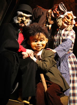 Scrooge sitting down with red-masked Bob Cratchit behind him leaning into Mrs. Cratchit with gold mask and blad dress and apron, and she has her hand on the shoulder of big-headed Tiny Tim who is sitting and leaning on Scrooge's arm