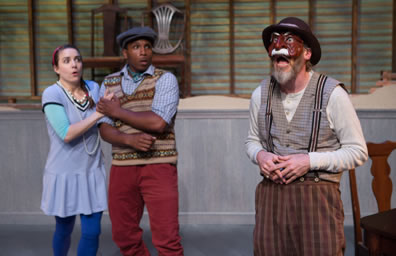 Harpagon in checkered pants, suspenders, gray checked vest, and dingy long-sleeved undershirt plus fodora hat and brown mask with a white mustache stands to the right while Elise in blue schoolgirl dress with white tights and Cleante in red pants, brown striped-patterned seater vest and blue shirt plus dog cam clasp each other in shock over what their father is saying. Chairs are on a platform in the background in front of a slatted wood wall.