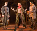 The Player with arms out and legs in a collosus stance talks with Guildenstern to his right, who is looking up and away, while Rosencrants on The Player's left looks at him. In the background is a stuffed chair behind Guildenstern and a attic ladder behind Rosencrantz and various junk.