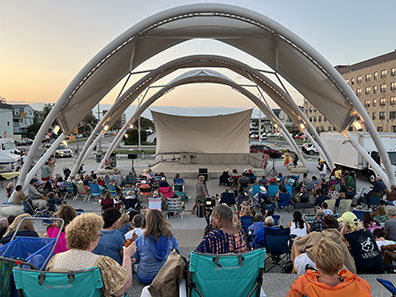 ROK-Amphitheater-Opening A view of the three-awning amphitheater looking from the boardwalk in toward the empty stage. People sitting in lawn chairs and on blankets, with buildings in the background and the sunset in the distance.