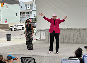 ROK-Midsummer-Theseus Theseus in red jacket, black pants, and pink hat and arms outspread, next to him is Hippolyta in camoflage pants and dark vest on a concreet stage with houses and a street intersection in. the background, the heads of people in lawn chairs in the foreground.