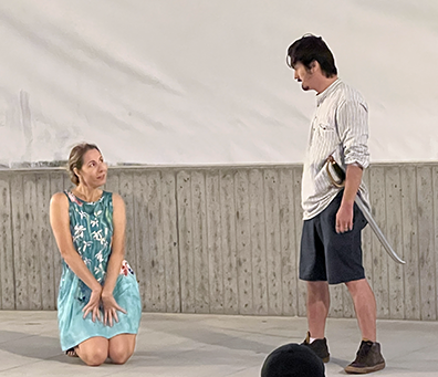 ROK-Midsummer_Spaniel Helena wearing an aqua sun dress with white blooms, kneels on the stage with her hands obediently pleading with Demetrius standing across from her wearing a kacki shirt, olive shorts, and a sword in his belt.