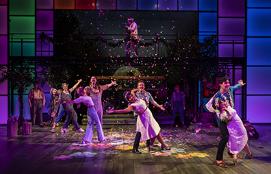 As You Like It, Shakespeare Theatre Company, Finali Dancers line up diagnally across stage at the end of their dance, a partner hanging on the arm of the other. Psychedelic lights on the floor, confetti falling, Hyman playing guitar on the gangway above.