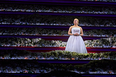 STC Evita-Inauguration Under a spotlight, Eva Peron wearing a bared-shoulder, white ball gown, sings amid layers of candlit fwhite flowers.