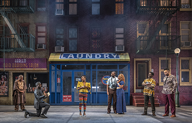 Characters in various West African-styled clothing (except the Pages, he in  common American short-sleeve shirt and pants with binoculars hanging around his neck from birdwatching, and she in floor length blue dress with small yellow X's) watch Ford's contritio in front of a street scene of the salon and laundryomat with tenements on either sice and a street lamp to one side. 